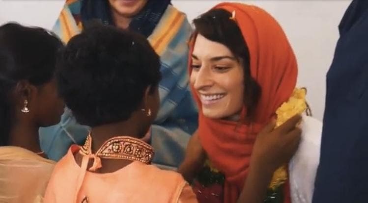 A woman in an orange scarf smiles as children in India welcome her with a marigold garland.